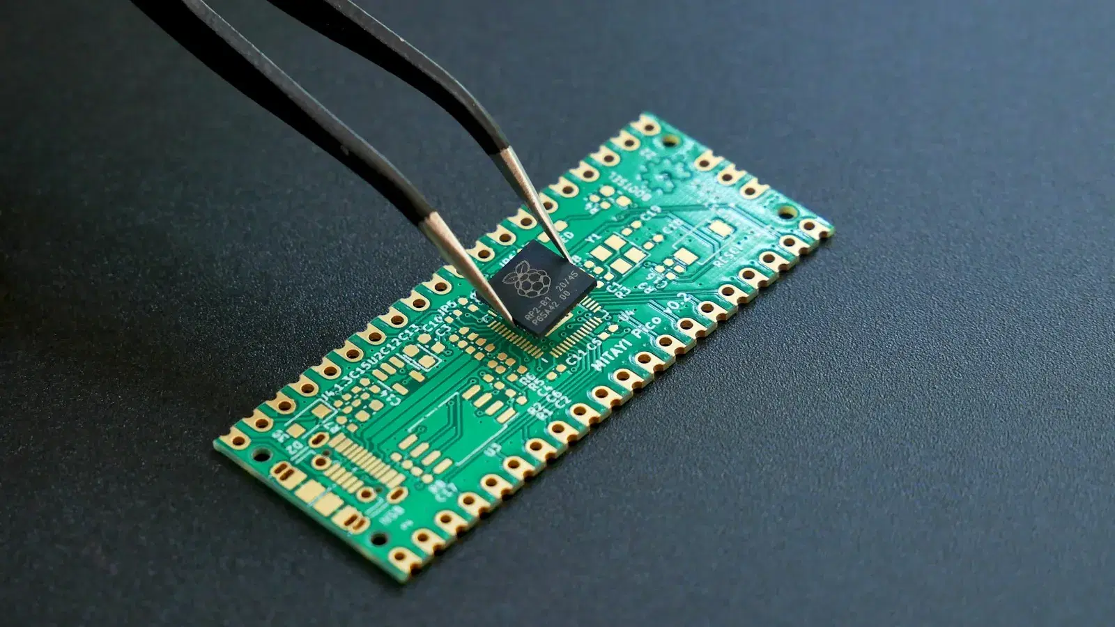 Tweezers placing a microcontroller chip onto a green printed circuit board.