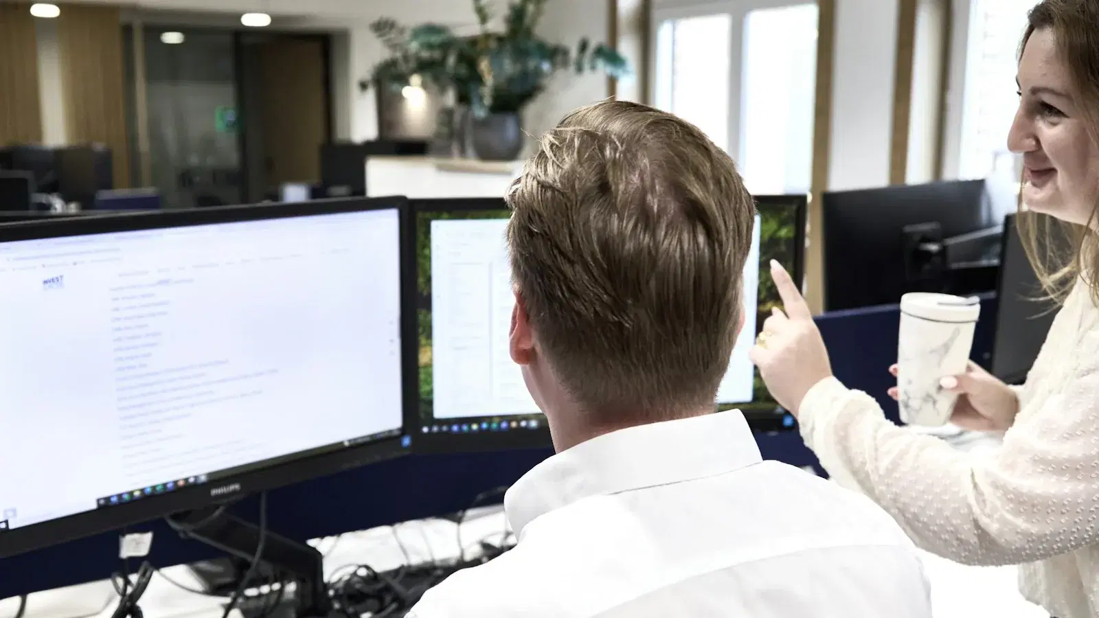 Two colleagues at a desk of monitors, one pointing something out on a screen to the other.