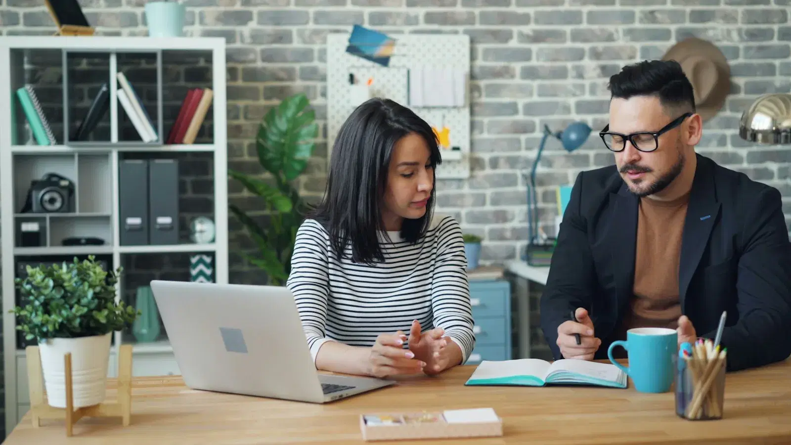 Two people going through work together at a desk, with a laptop and an open notebook between them.