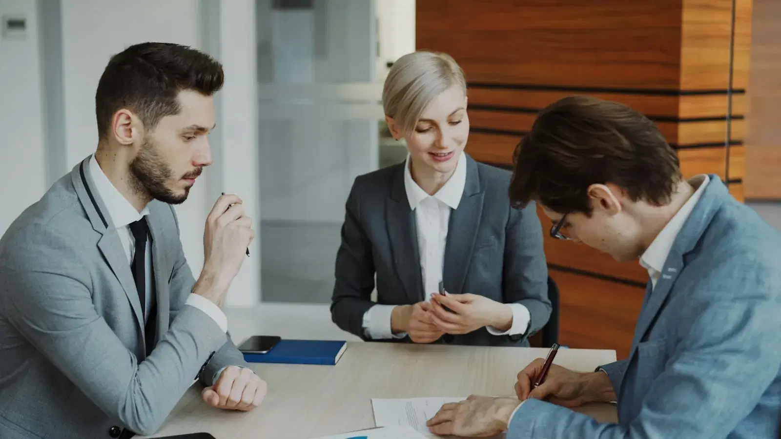 Two people signing a contract across a table while a third explains the terms.