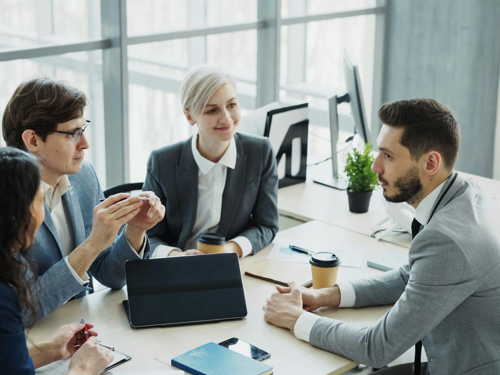 Four colleagues around a meeting-room table with a tablet and notes, mid-discussion